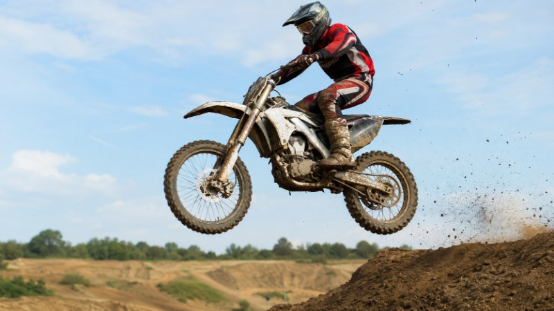 Jezdec v plné motokrosové výstroji skáče na motorce bez SPZ přes hliněný hrb na uzavřené terénní trati