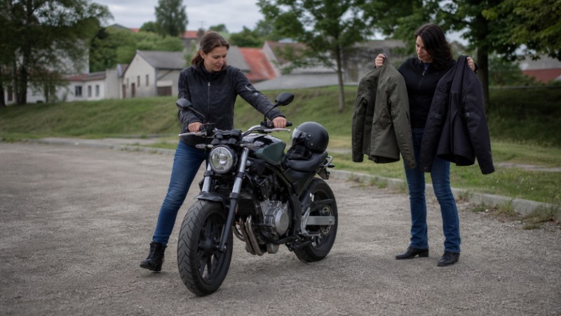 Motorkářka v přilbě a moto bundě stojí u zaparkované motorky a kontroluje posed v sedle