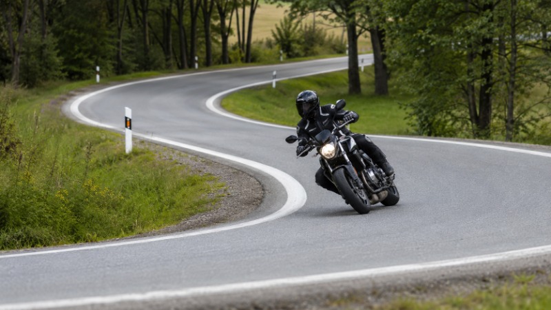 Motorkář v náklonu projíždí zatáčku na okresní silnici, pohled směřuje do oblouku